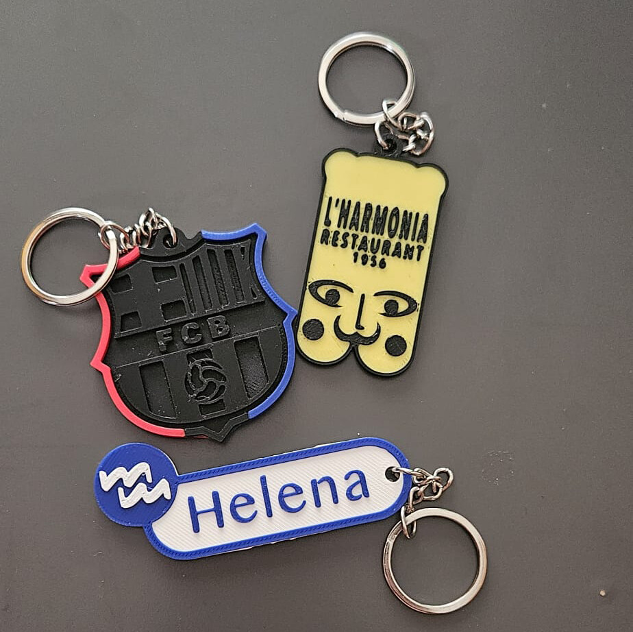 Tres llaveros impresos en 3D montados con argolla y cadena: escudo del FCB, L'Harmonia Restaurant 1956 y nombre Helena con logo del hospital